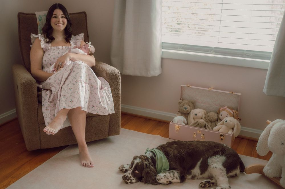 New mother in floral dress holds newborn in nursery chair while spotted dog rests on floor nearby.