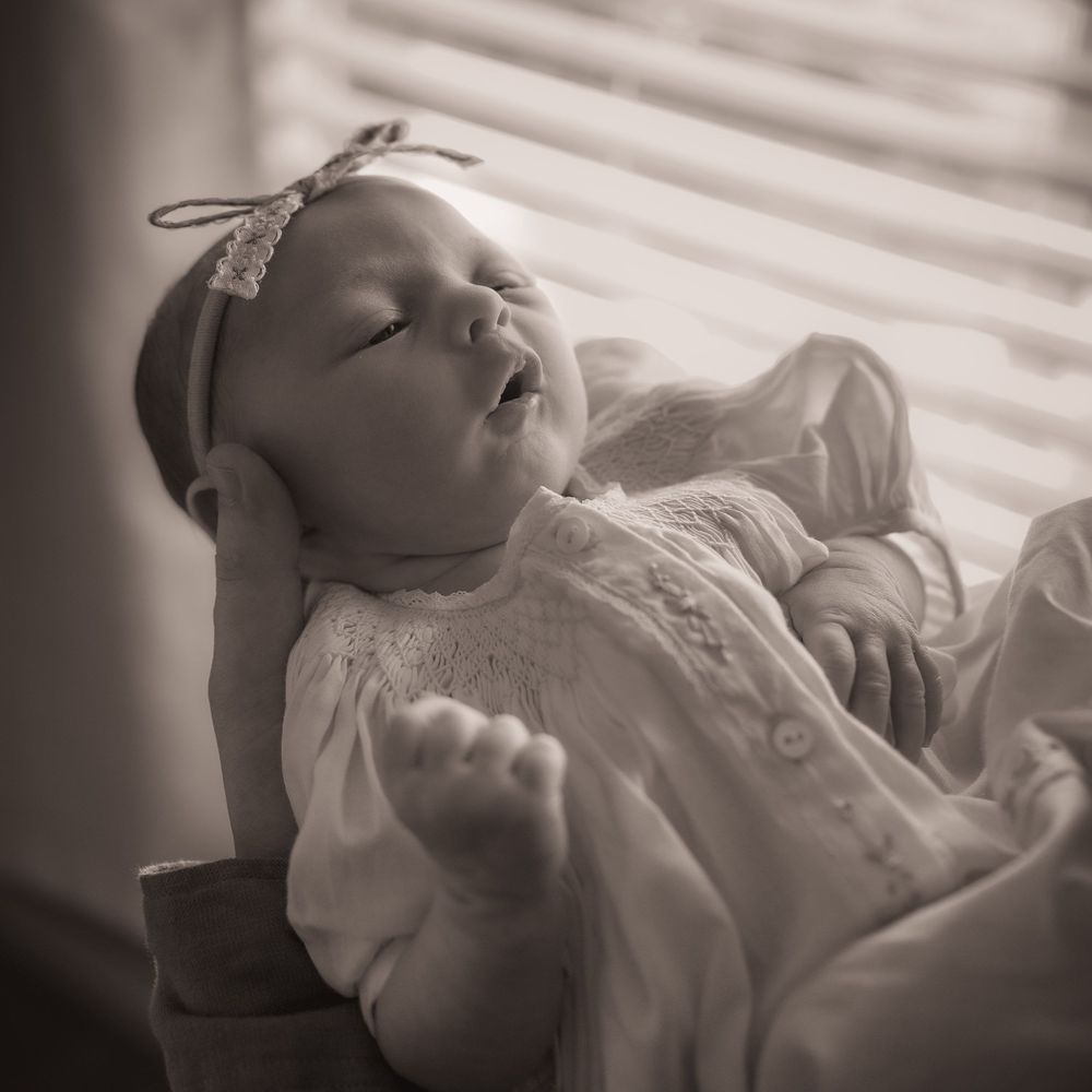 Black and white photo of a sleeping newborn baby girl wearing a white outfit and lace headband, bathed in soft window light.