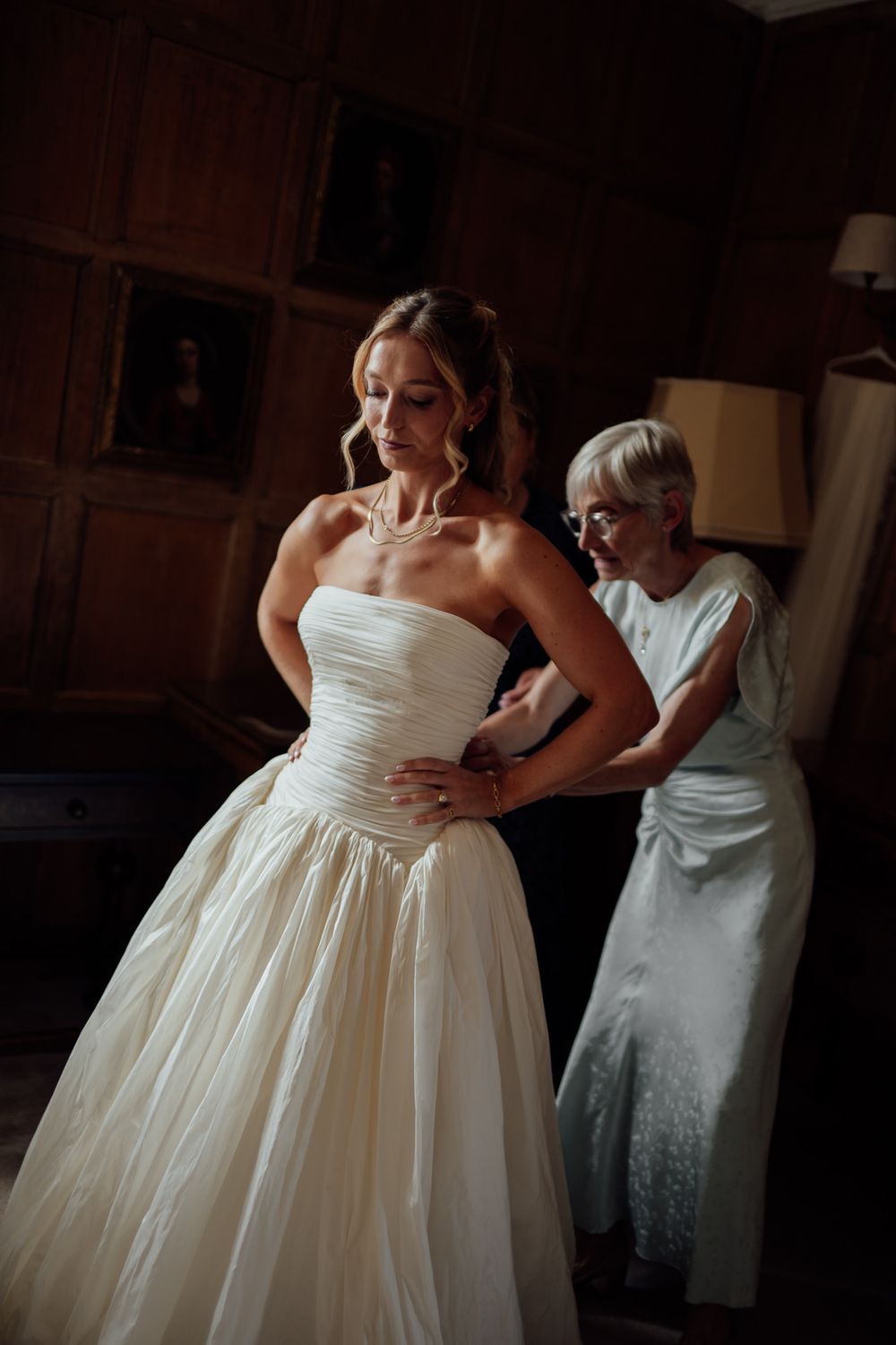 Bride in strapless white ball gown being helped by older woman in elegant wood-paneled room.
