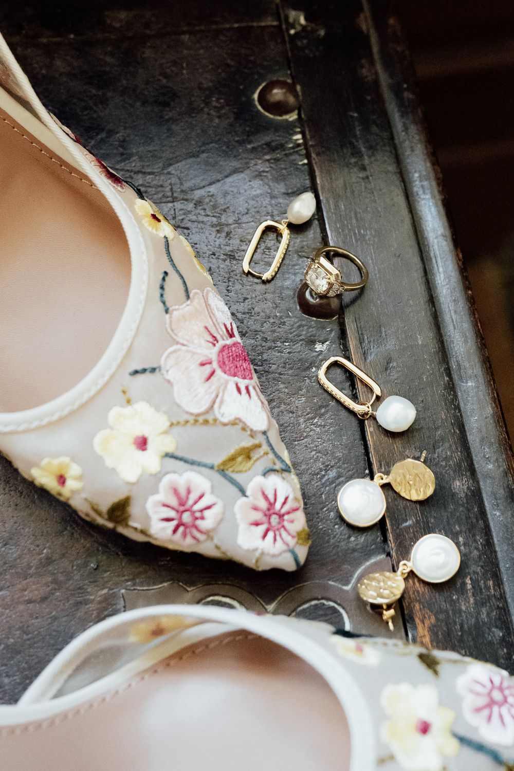 Floral embroidered bridal heels beside gold and pearl drop earrings on a rustic wooden surface.