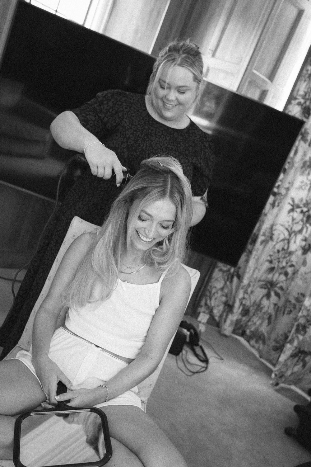 Black and white photo of a smiling woman getting her hair styled by a hairdresser in a bright room.