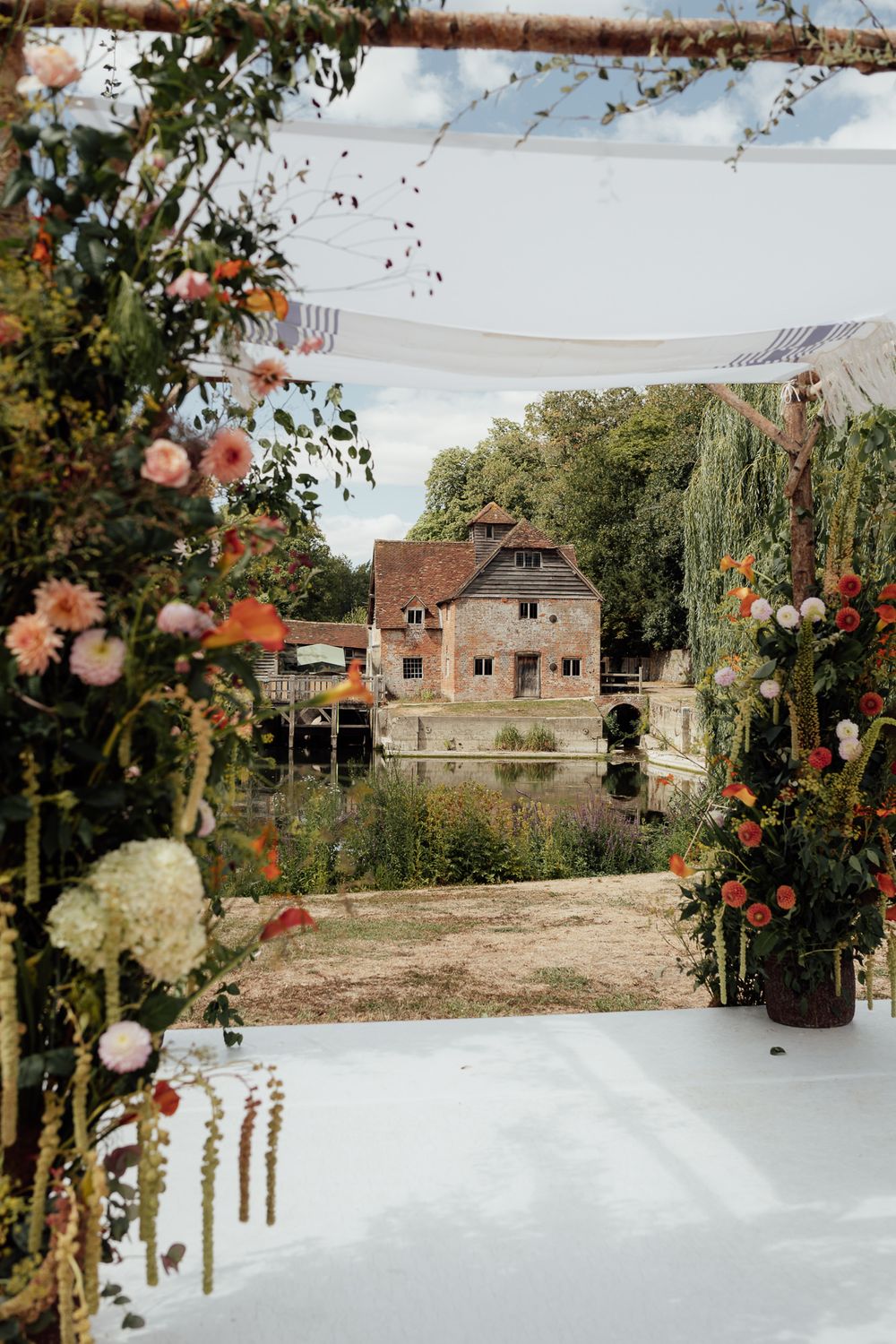Floral arch frames a rustic stone mill beside a tranquil river, surrounded by lush greenery at an outdoor wedding.