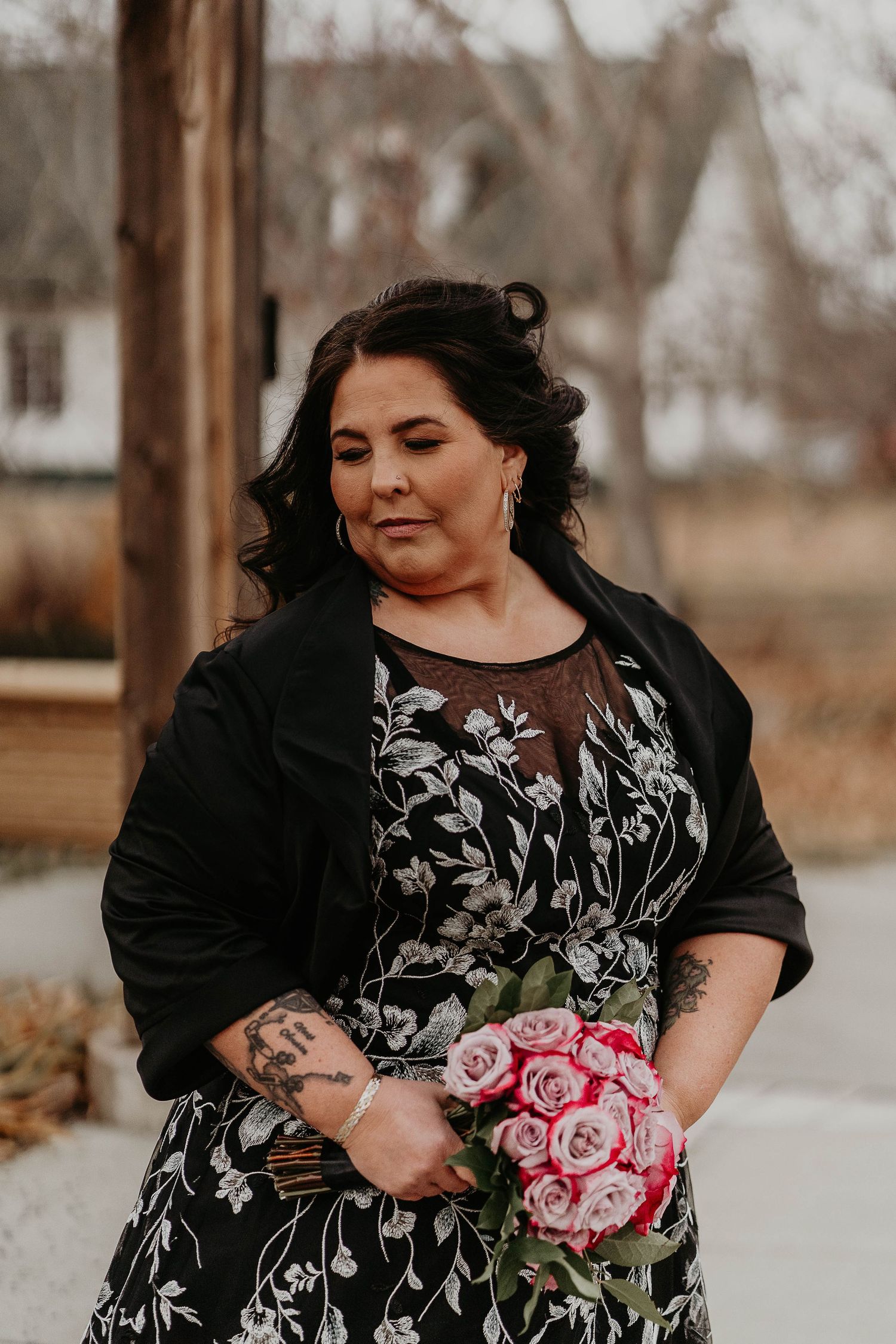 Woman in floral dress and black blazer holds pink rose bouquet, featuring tattoos, at outdoor wedding venue.