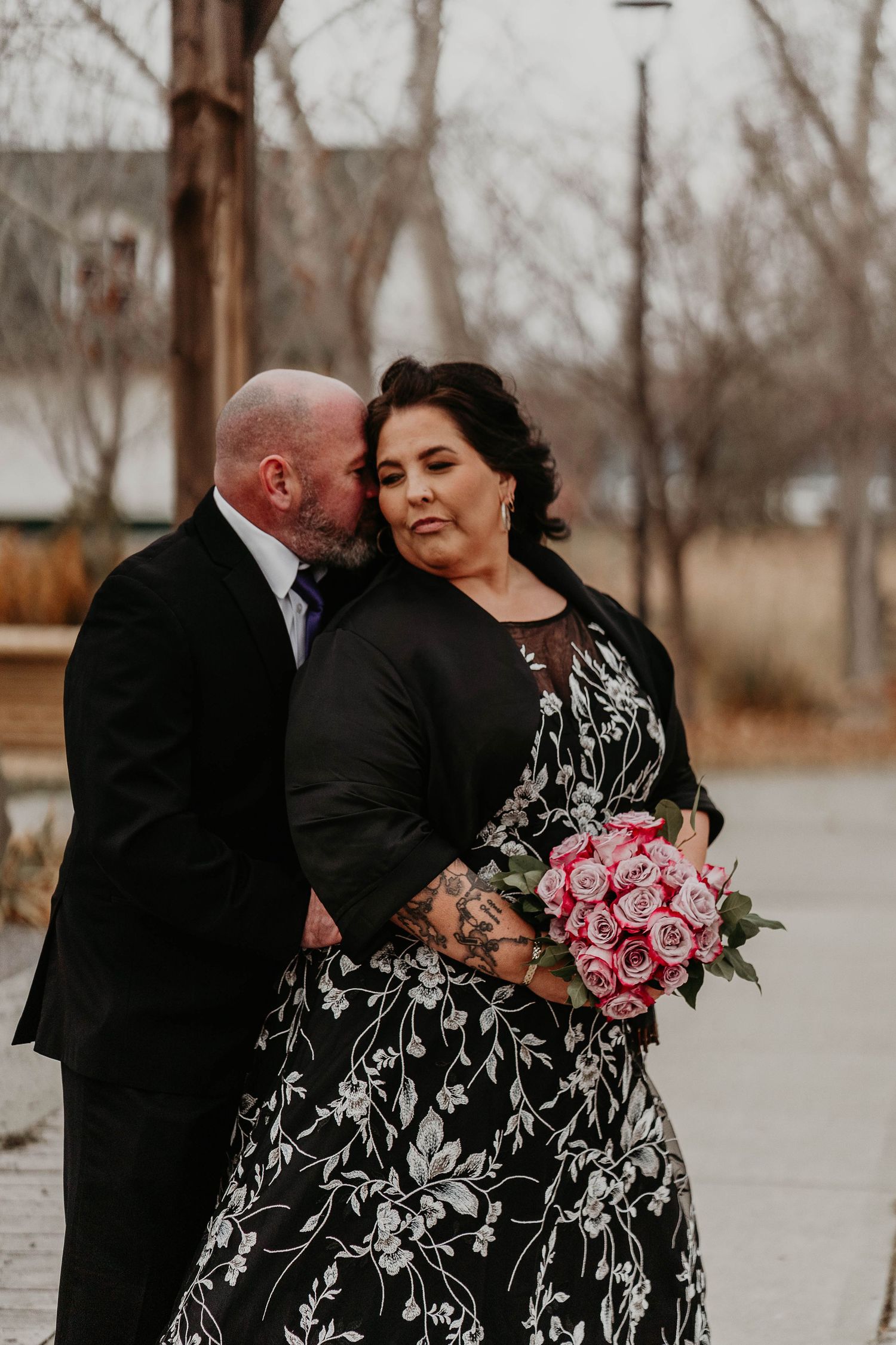 Couple in wedding attire, groom kissing bride's cheek, bride holding pink rose bouquet, outdoors.