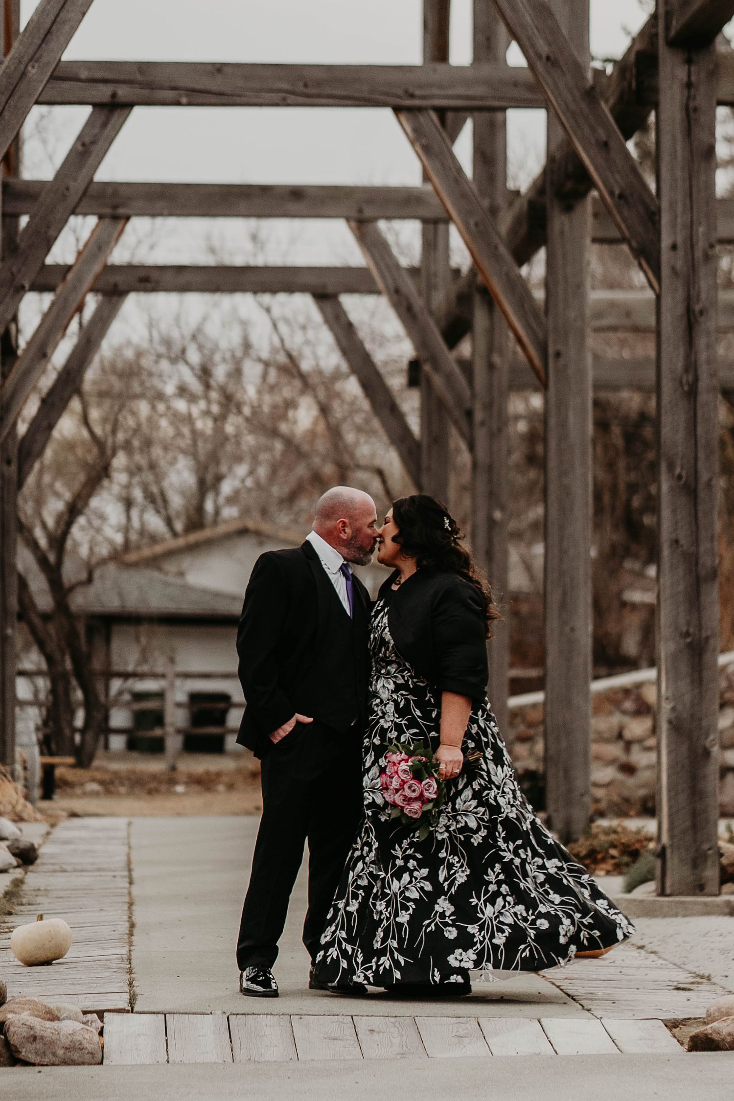 Couple sharing a romantic kiss under rustic wooden pergola, bride in black floral gown holding pink bouquet.