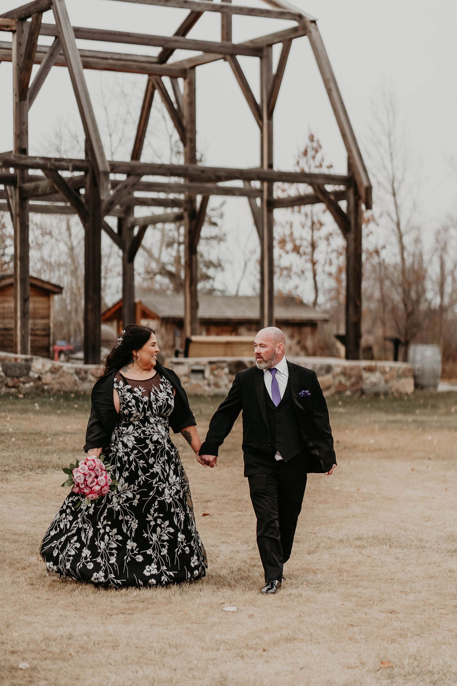 Bride in black floral gown holds bouquet walking with groom in black suit at rustic outdoor wedding venue.