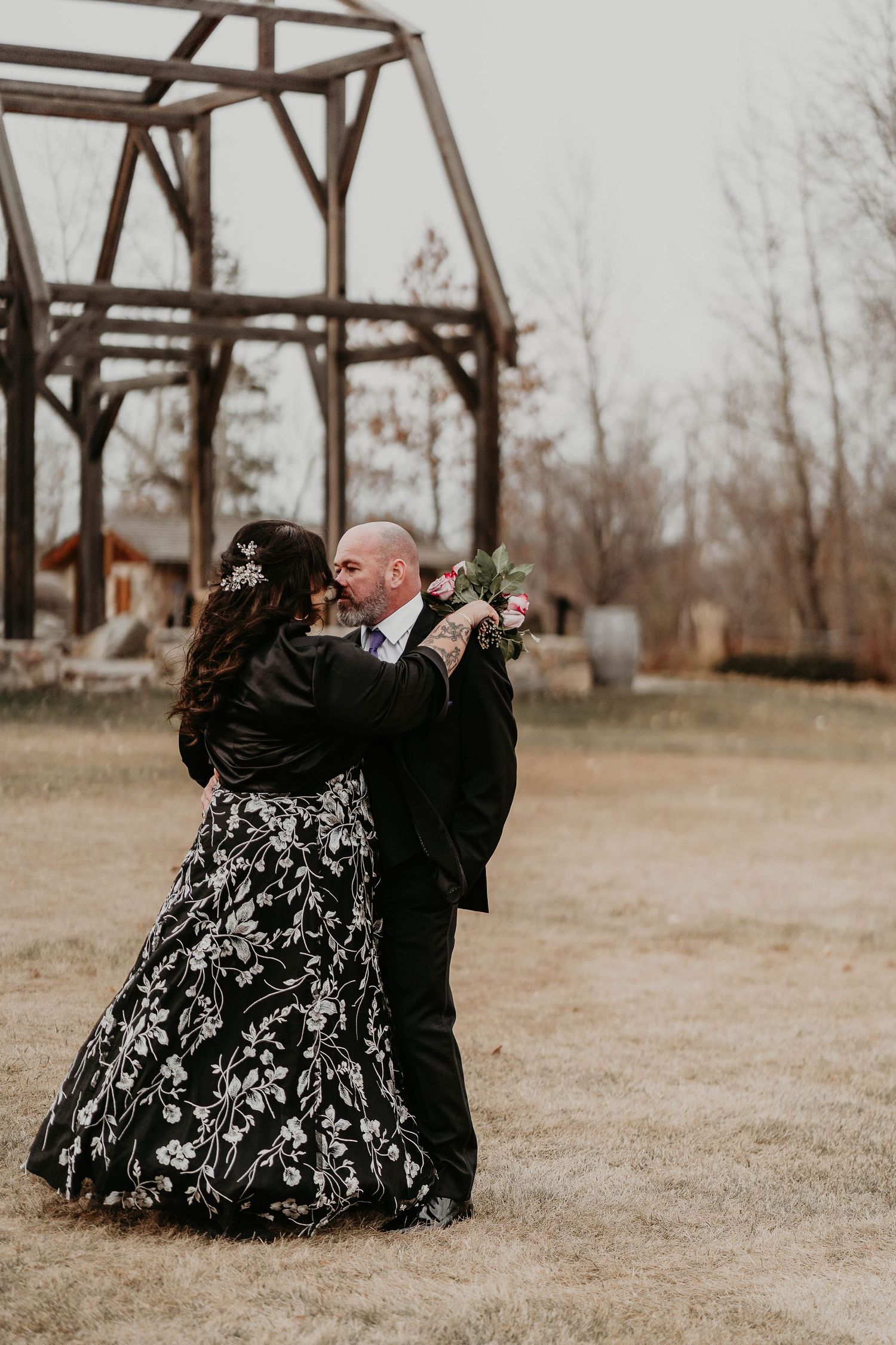 Bride and groom share romantic embrace in black wedding attire before rustic wooden structure at outdoor ceremony.