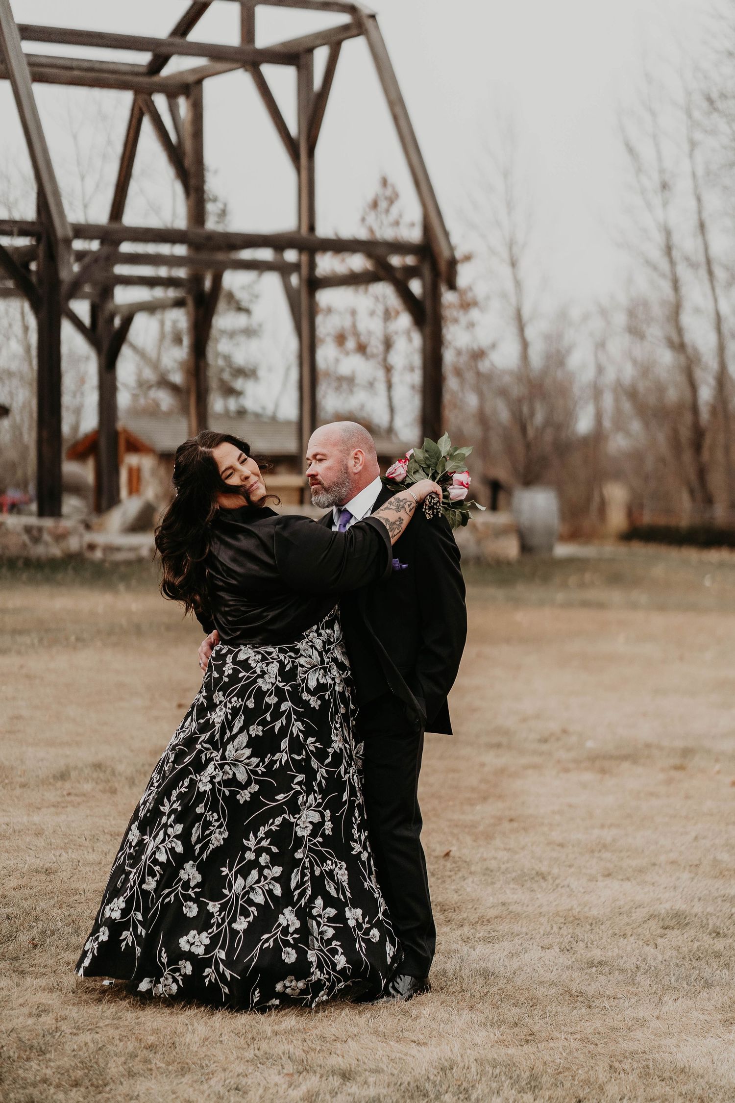 Couple embracing outdoors in rustic wedding setting, bride in black floral gown holding pink bouquet, groom in black suit.
