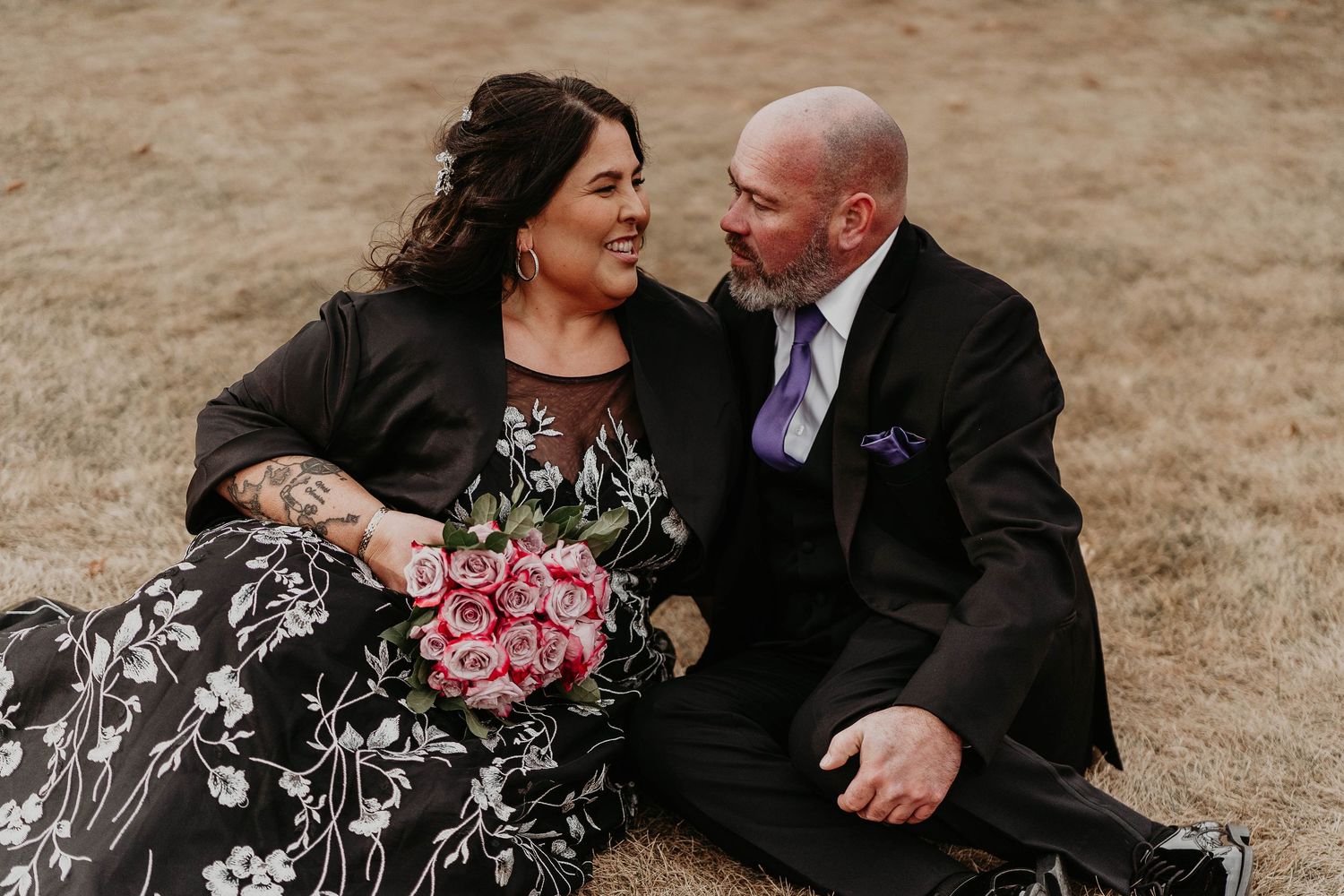 A couple in wedding attire sits on grass, smiling at each other, bride holding pink roses bouquet.