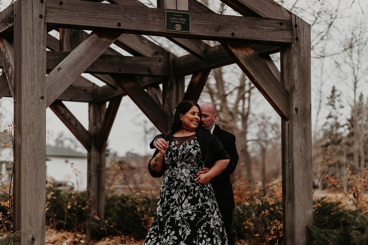 Couple posing under a rustic wooden pergola in a garden, woman in floral dress, man in black suit.