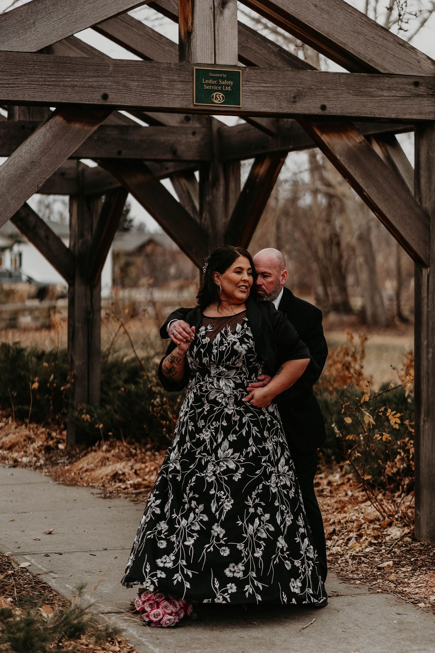 Couple embracing under rustic wooden pergola, bride in black floral gown, groom in suit, outdoor autumn wedding portrait.