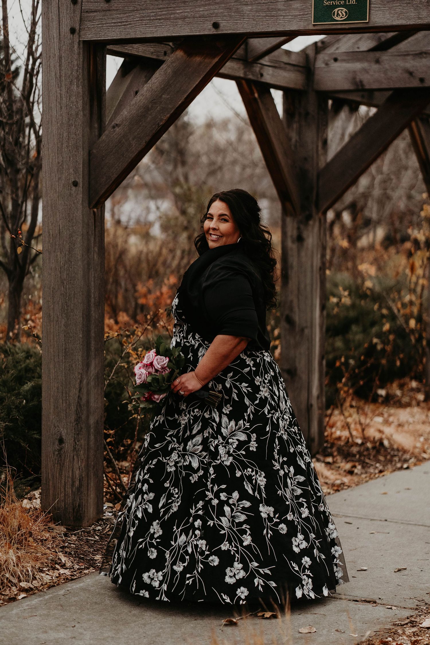 Woman in black floral ball gown holding pink bouquet smiling under wooden pergola in autumn garden setting.
