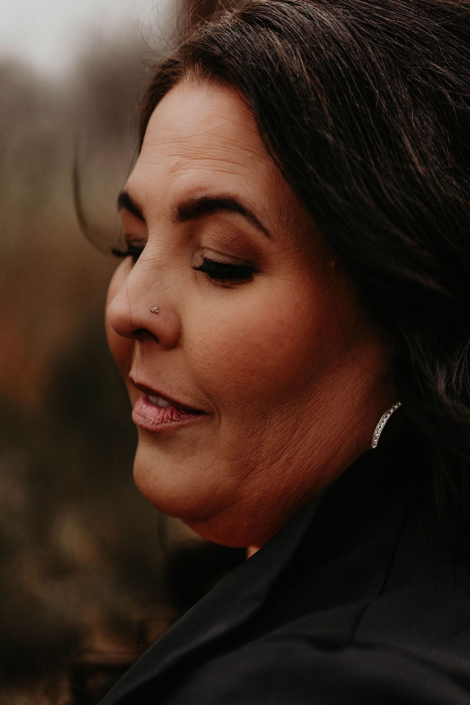 Close-up portrait of a woman with dark hair, nose piercing, and diamond earrings, eyes closed, wearing a black jacket.