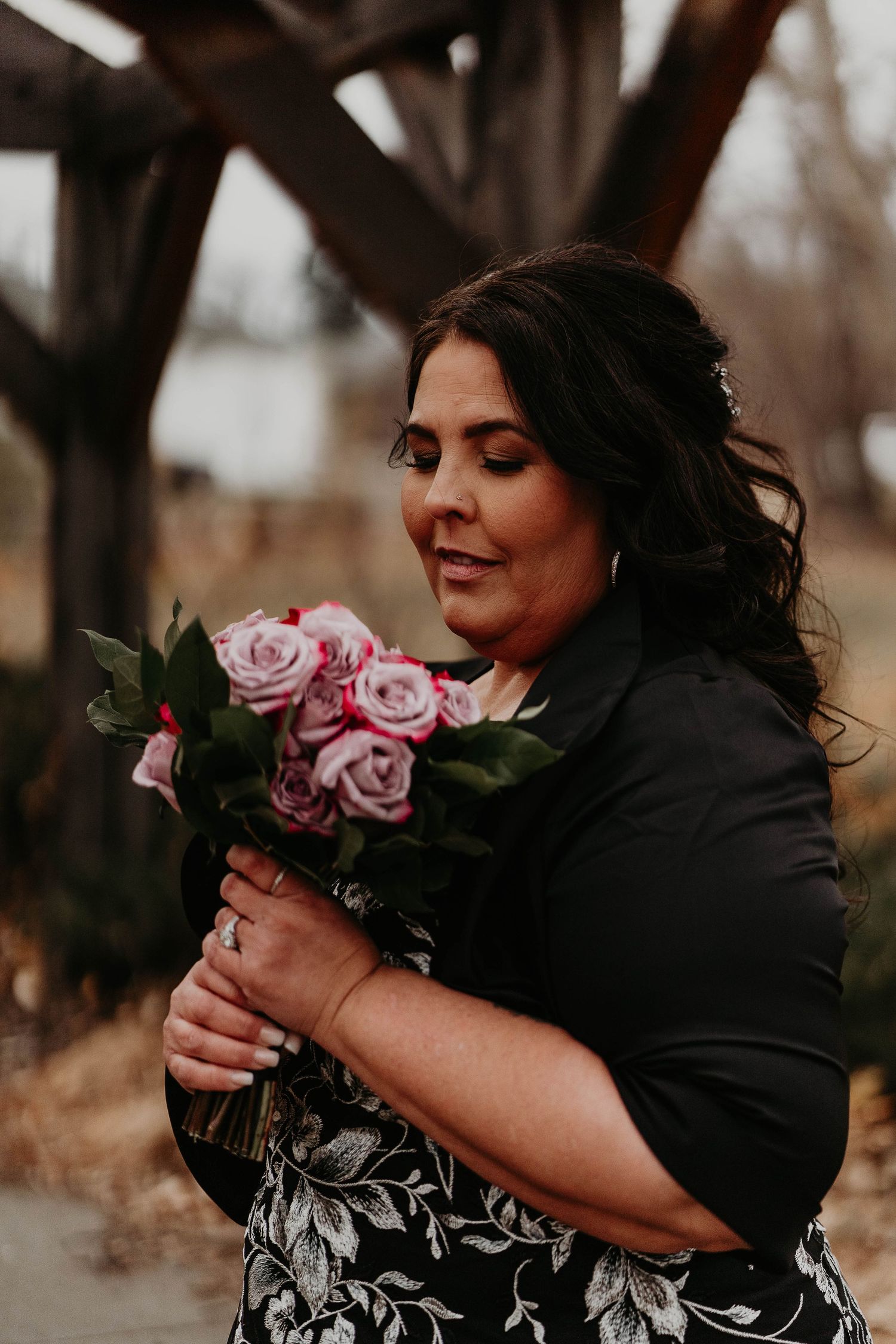 Woman in black jacket holding pink roses bouquet outdoors near wooden structure, smiling softly downward.