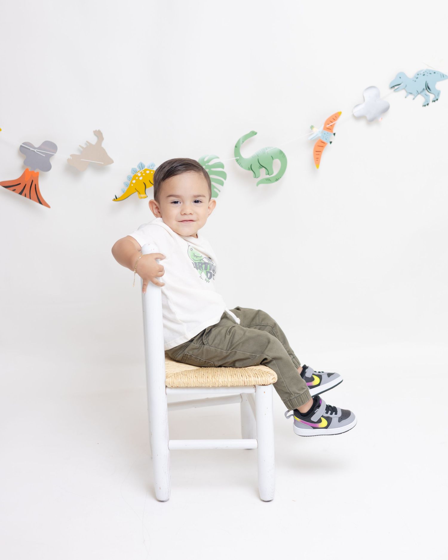 Young toddler boy sitting on white chair wearing white tee and olive pants with dinosaur banner backdrop.