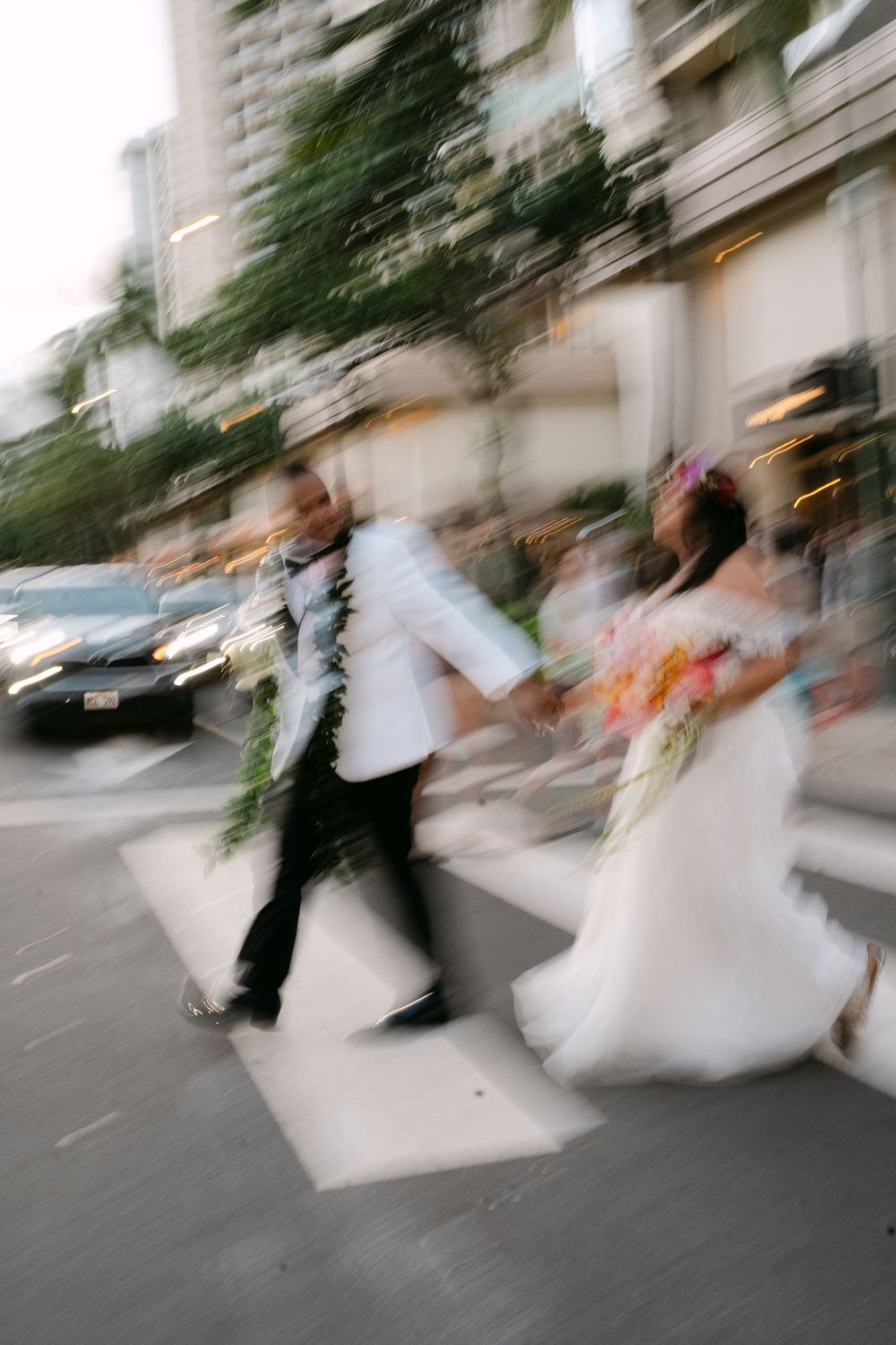 Blurred motion photo of a bride and groom crossing a street crosswalk in wedding attire with colorful bouquet.