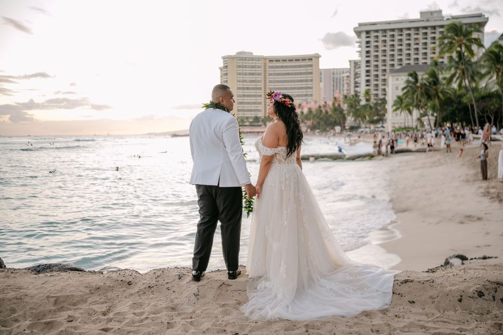 Bride and groom holding hands on Waikiki Beach at sunset, with palm trees and hotel in background.