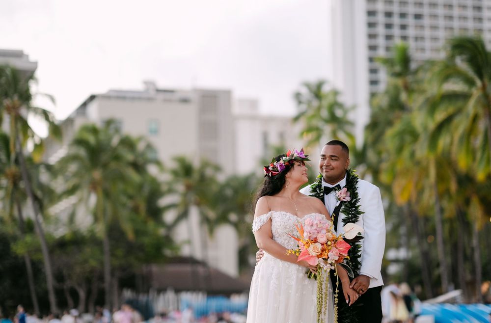 Bride and groom pose on Waikiki Beach, Hawaii, with palm trees and hotels in the background on their wedding day.