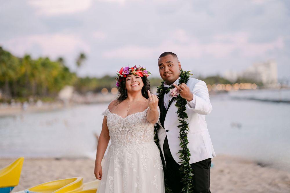 Newlywed couple posing on Waikiki Beach, bride in floral crown and gown, groom in white jacket with green lei.