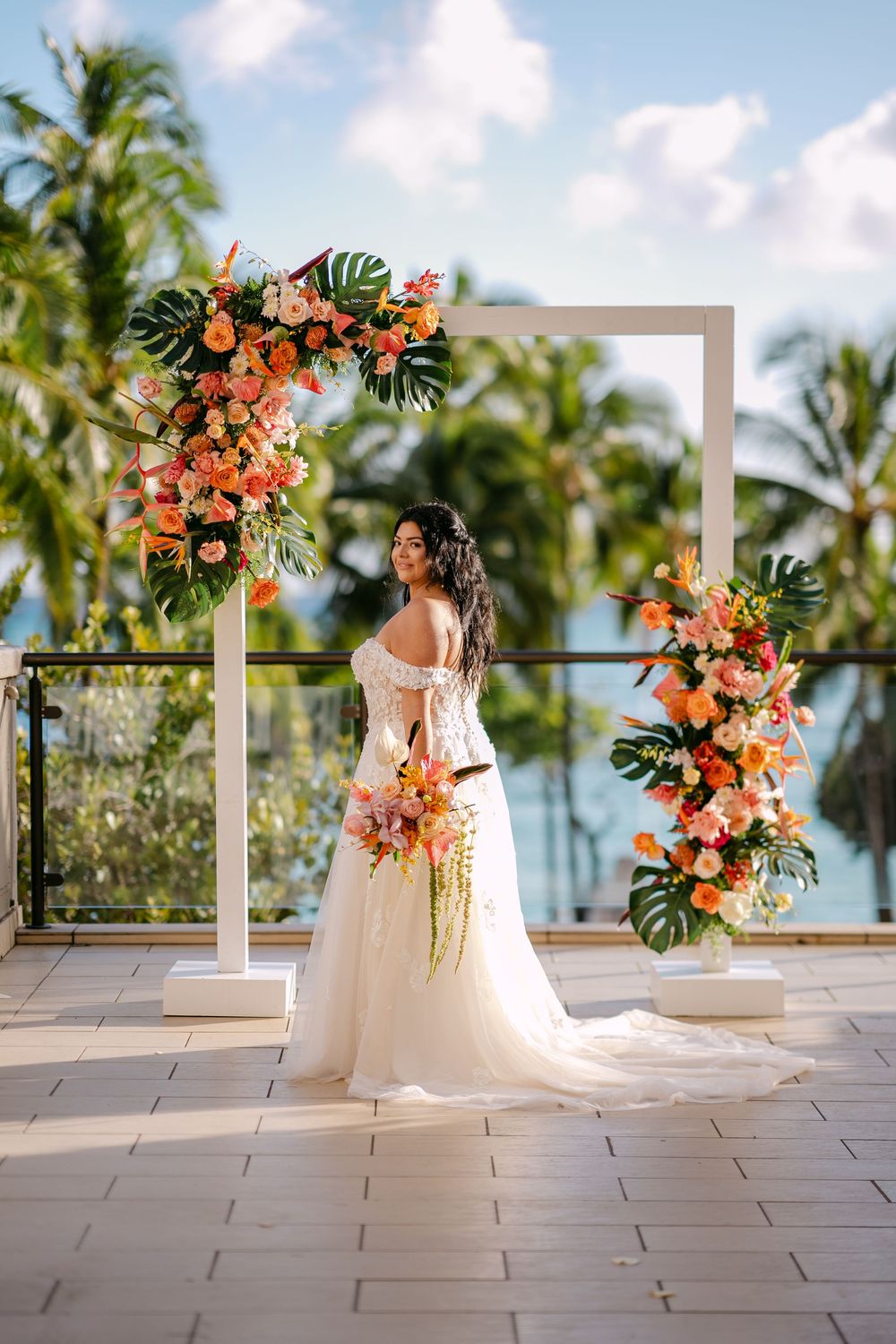 Bride in white off-shoulder gown holds tropical bouquet before floral arch with orange blooms and palms in Hawaii.