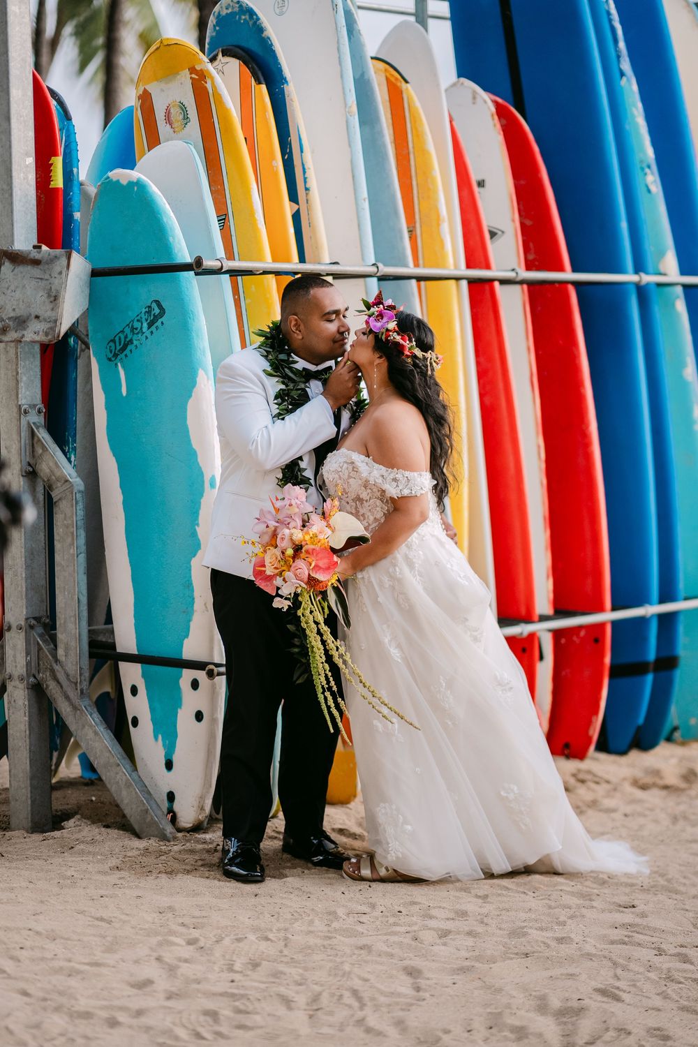 Bride and groom share a romantic moment on the beach in front of colorful surfboards in Hawaii.