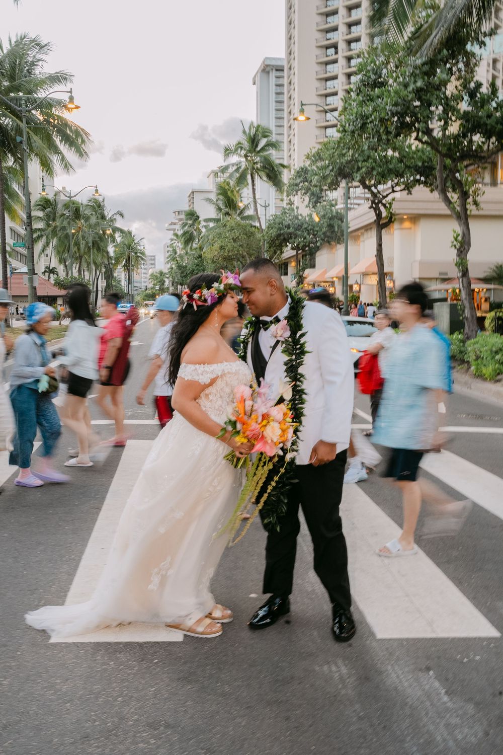 Bride and groom kissing on a crosswalk in Waikiki, Hawaii, with blurred pedestrians and palm trees in the background.