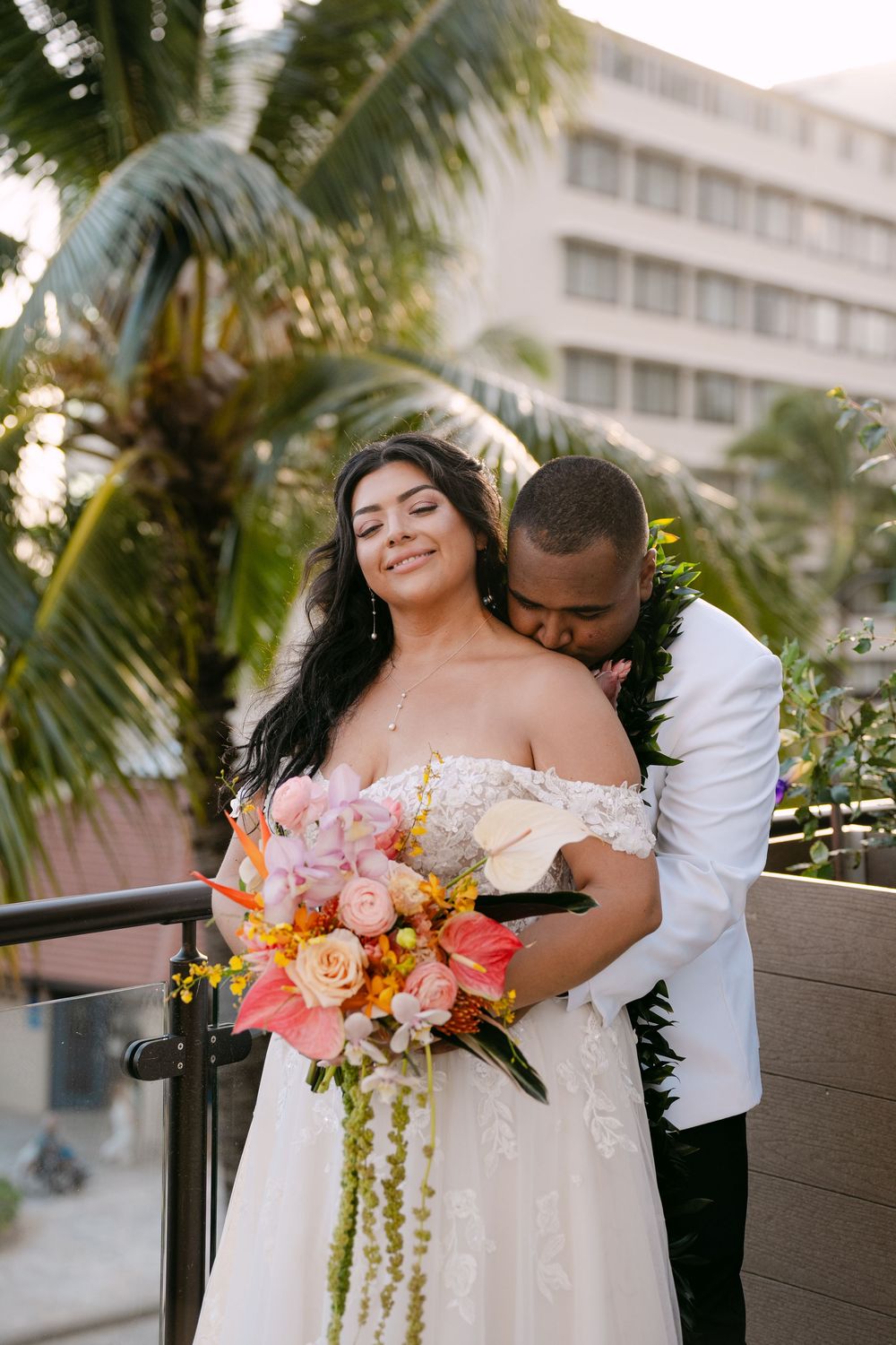 A groom in a white suit kisses his bride's neck as she smiles, holding a tropical bouquet on a balcony.