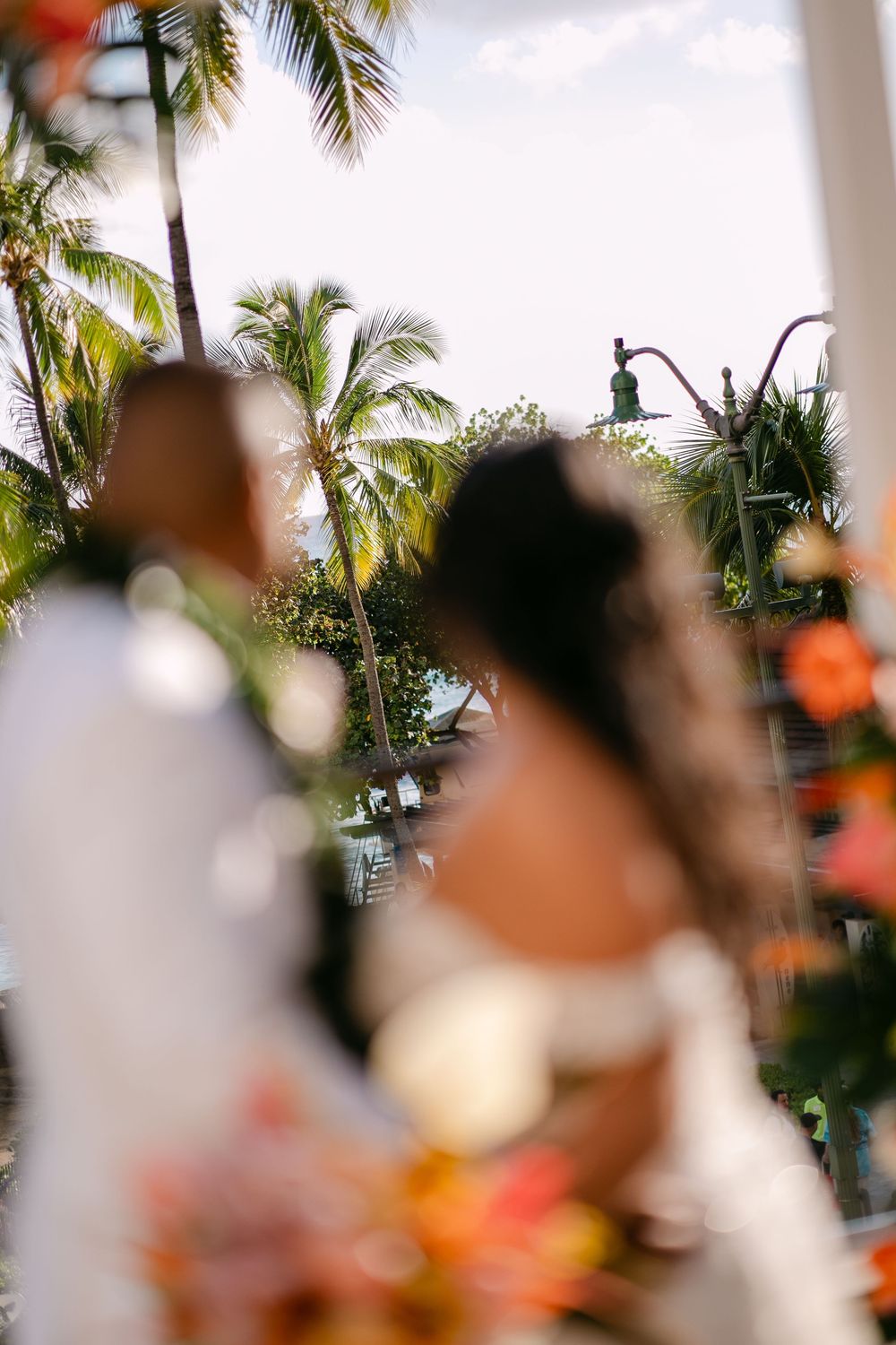 Blurred foreground of a tropical wedding with palm trees, colorful flowers, and a couple in formal attire outdoors.