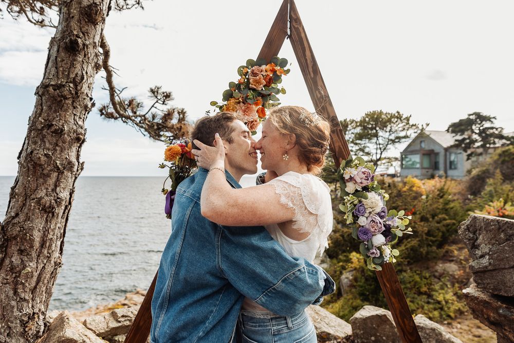 Two brides share a romantic kiss under a floral arch by the ocean at their outdoor wedding ceremony.