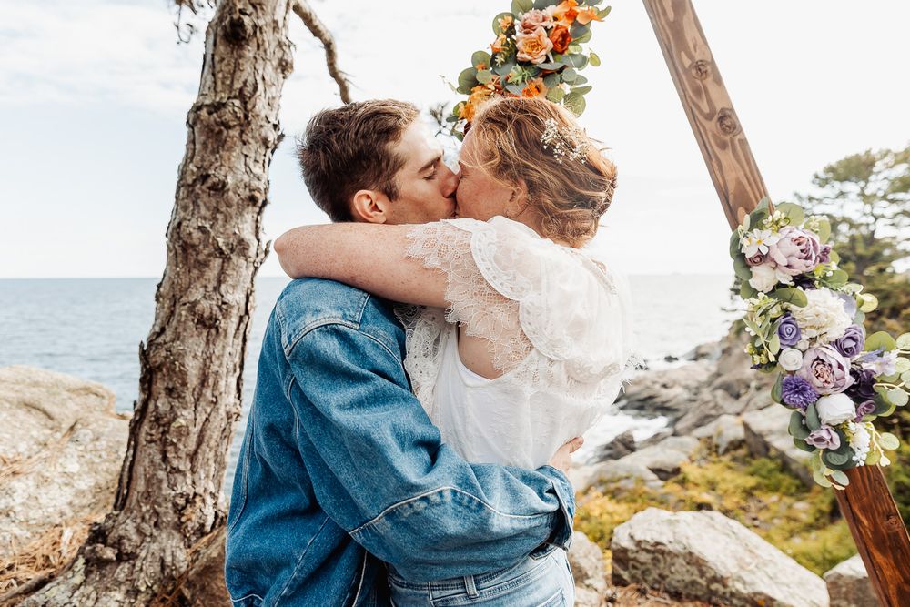 Couple sharing a romantic kiss at outdoor wedding ceremony by the sea, framed by wooden arch with colorful flowers.