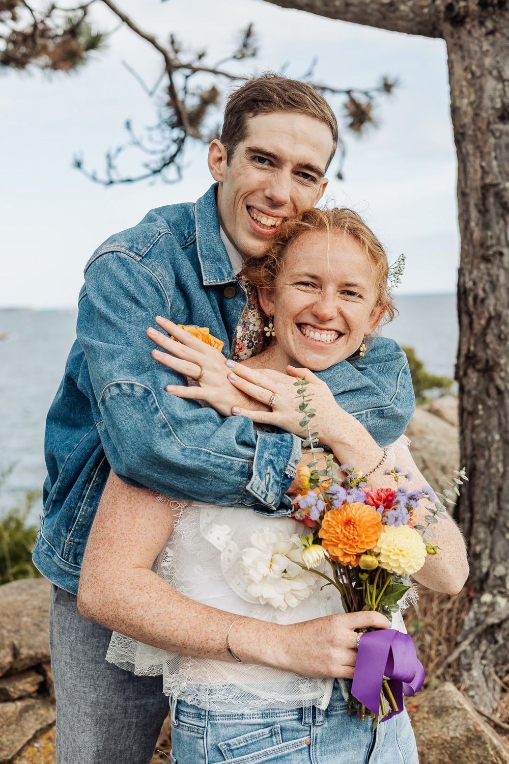 Happy couple embracing outdoors near the ocean, woman holding colorful bouquet with purple ribbon, both smiling.