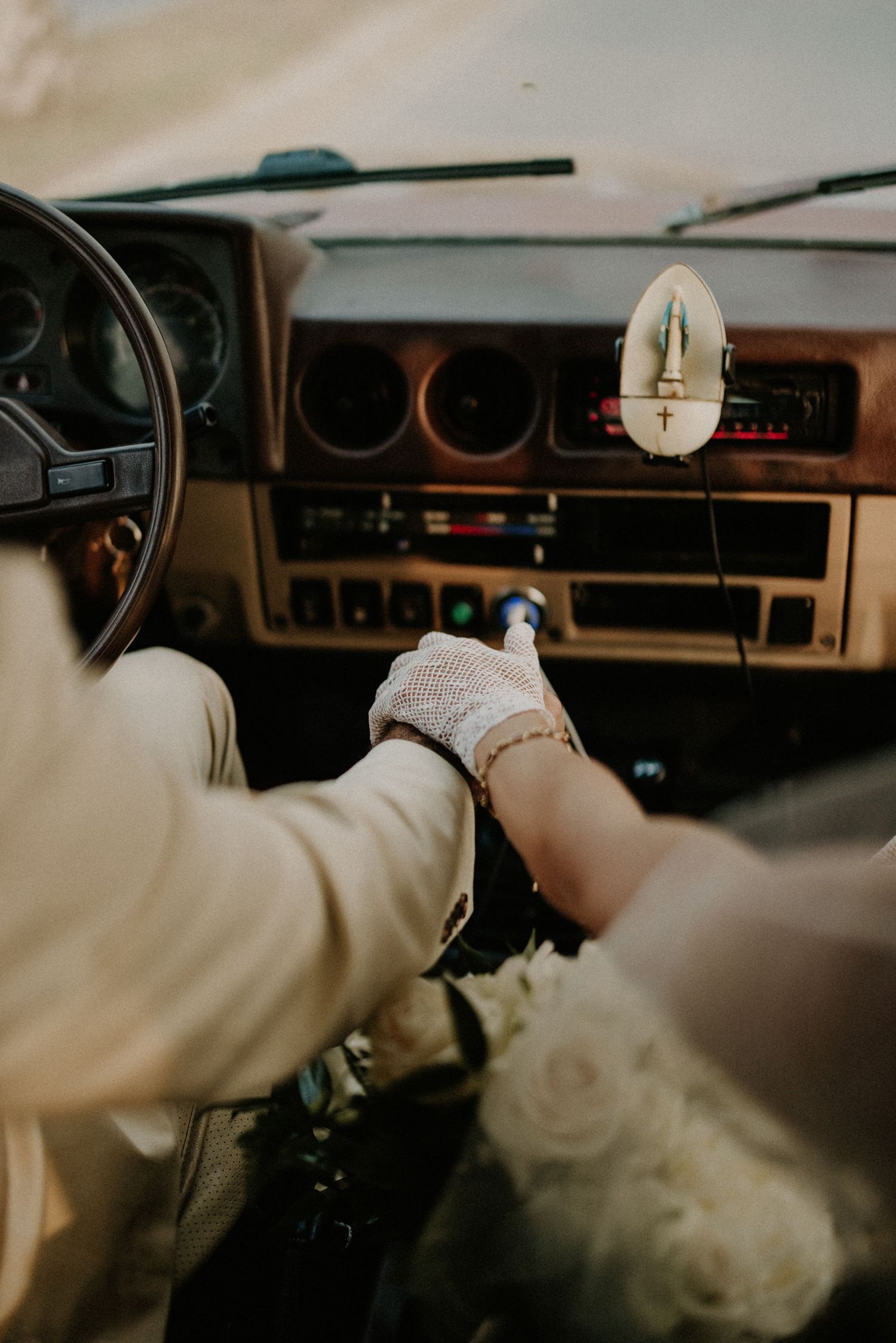 Bride and groom holding hands in a vintage car interior with white roses and lace gloves.