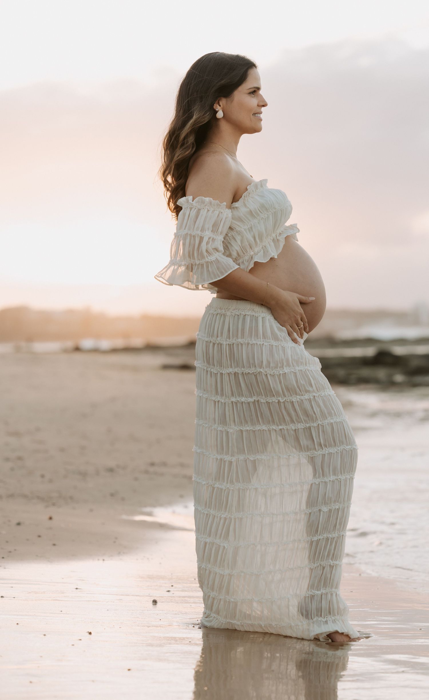 Pregnant woman in white off-shoulder dress standing on beach at sunset, cradling her baby bump.