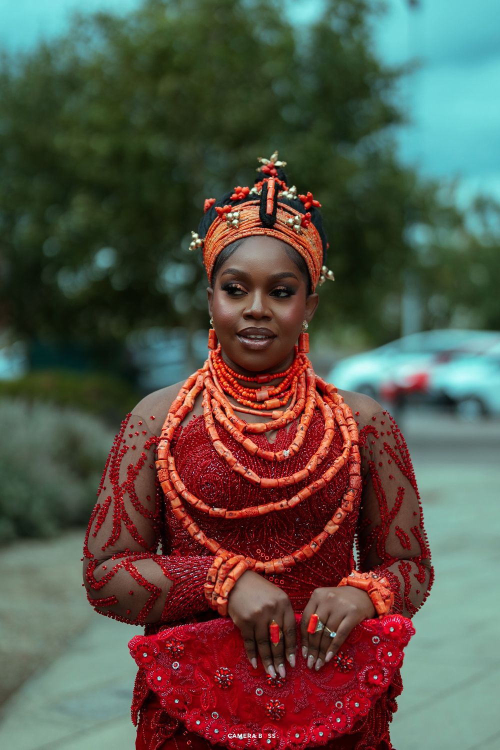 Woman in traditional Nigerian Edo bridal attire with coral beads, red lace dress, and ornate headdress outdoors.