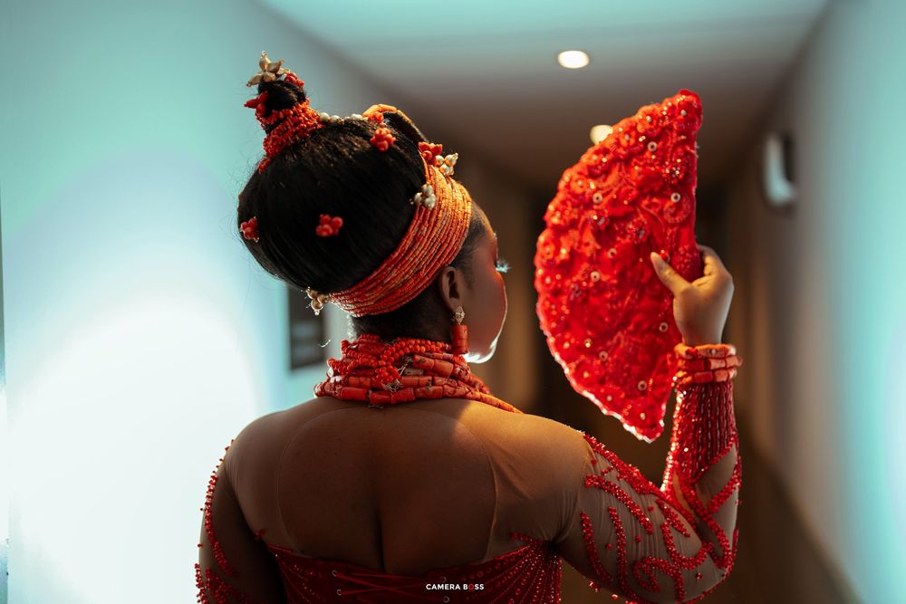 A woman in traditional red Nigerian bridal attire holds a red floral fan, adorned with coral beads and headpiece.