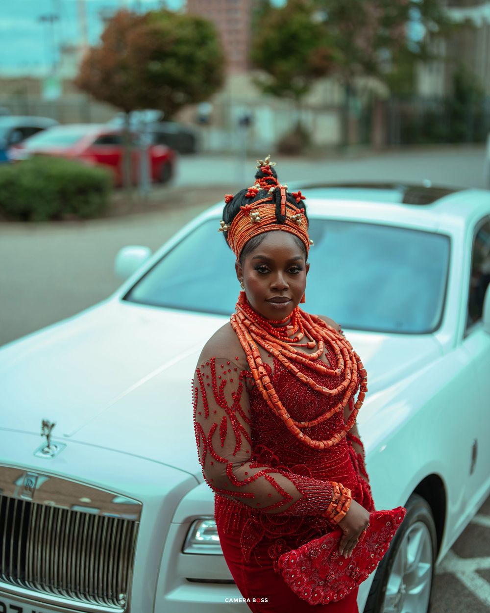 Woman in traditional Nigerian Edo bridal attire with coral beads and headpiece posing beside a white Rolls-Royce.