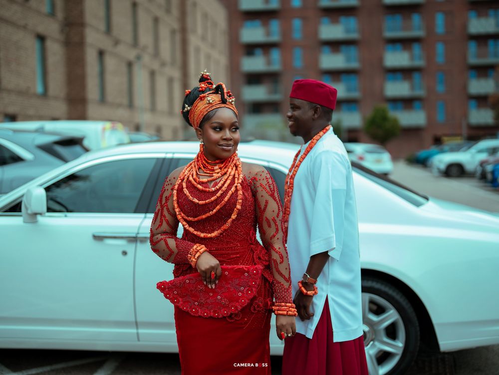 Nigerian couple in traditional red Igbo attire posing beside a white car outdoors.