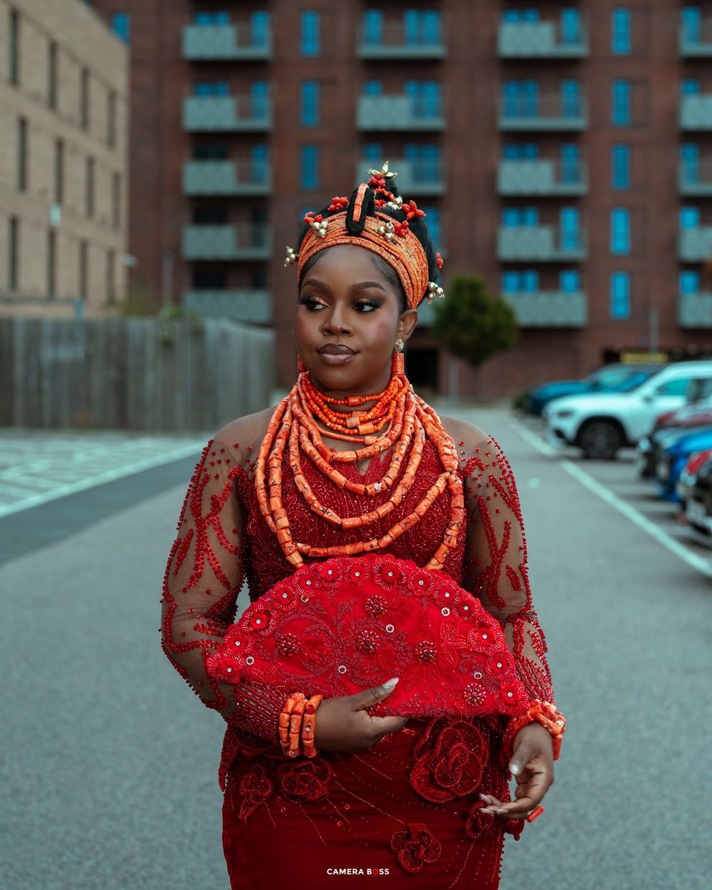 Woman in traditional Nigerian Urhobo red coral beaded attire and headdress holding a decorative fan outdoors.