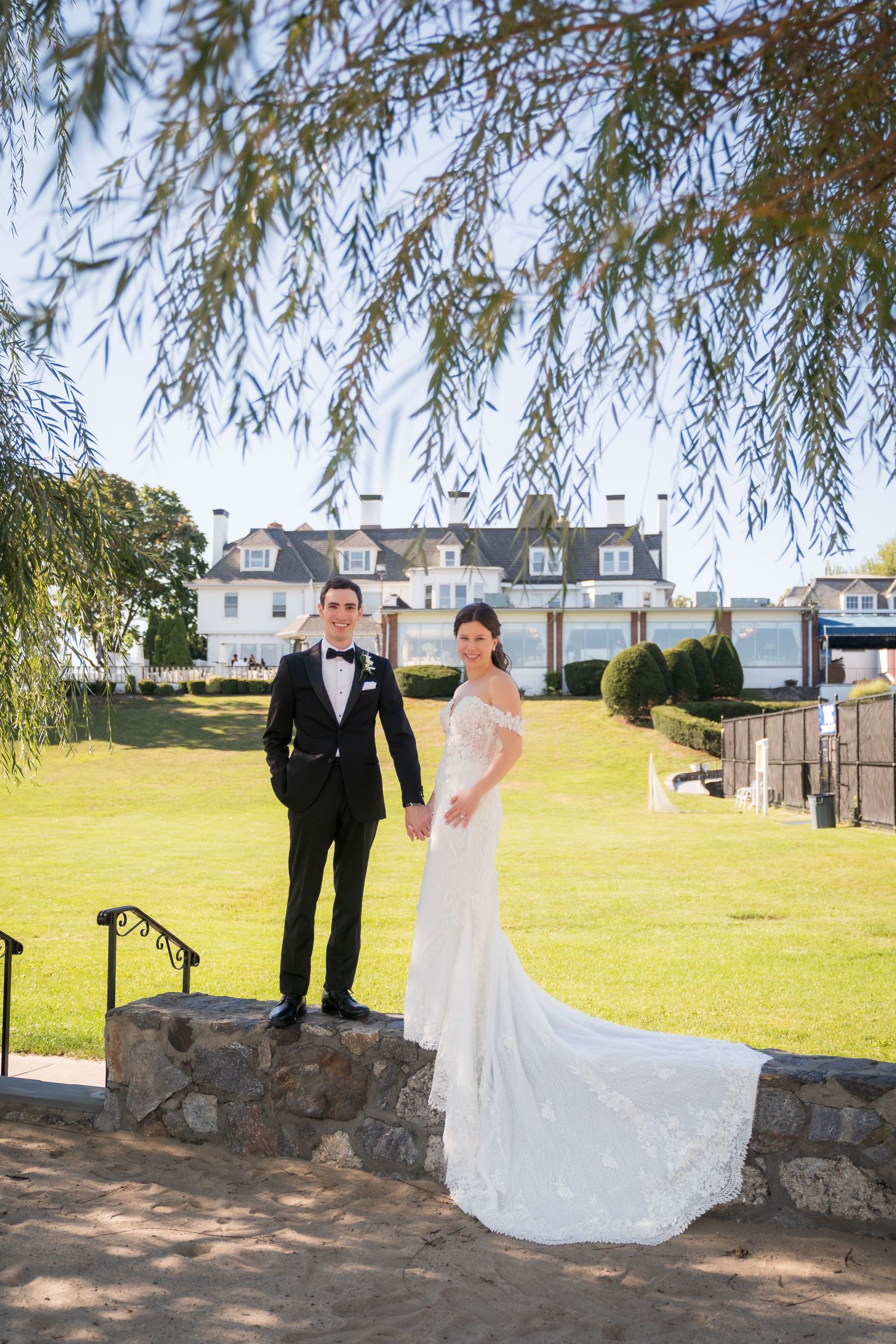 Bride in lace gown and groom in tuxedo pose on stone wall with elegant estate and green lawn backdrop.