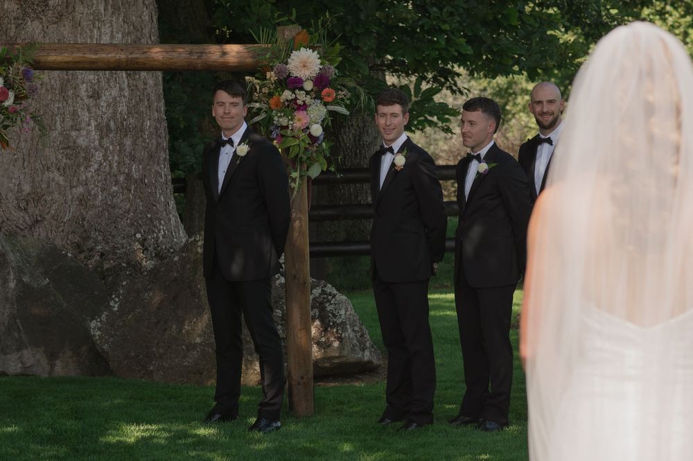 Groom and groomsmen in black tuxedos await bride in white veil at outdoor wedding ceremony near floral arrangement.