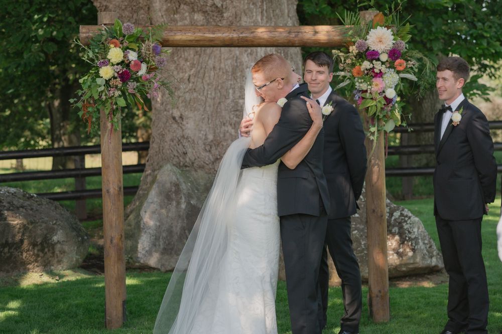 Bride and groom share first kiss under rustic wooden arch adorned with colorful flowers at outdoor wedding ceremony.