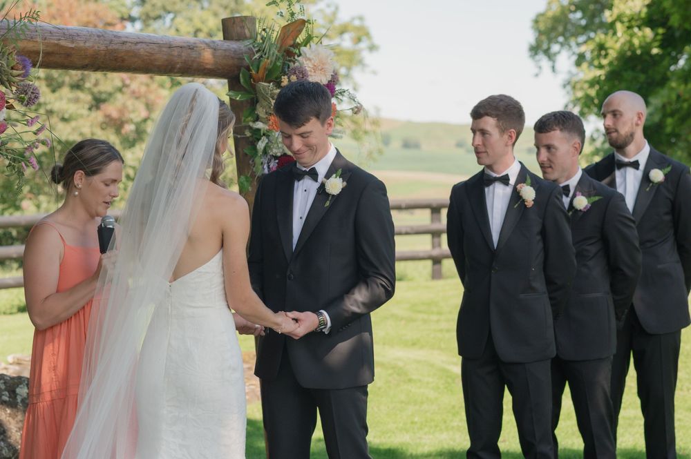 Outdoor wedding ceremony with groom in black tuxedo, groomsmen, and bridesmaid at rustic wooden arch.