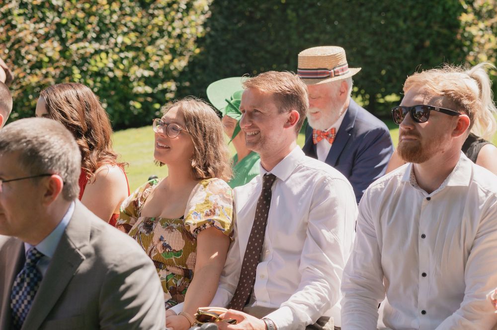Guests smiling and laughing outdoors at a sunny wedding ceremony, dressed in smart casual attire.