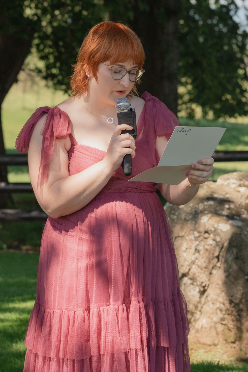 Red-haired woman in pink dress reads from paper while speaking into microphone outdoors at garden event.