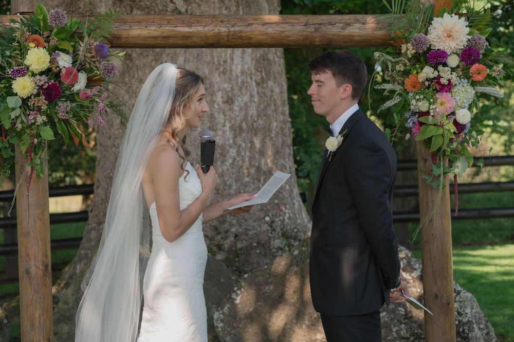 Bride reading vows to groom under rustic wooden arch adorned with colorful flowers during outdoor wedding ceremony.
