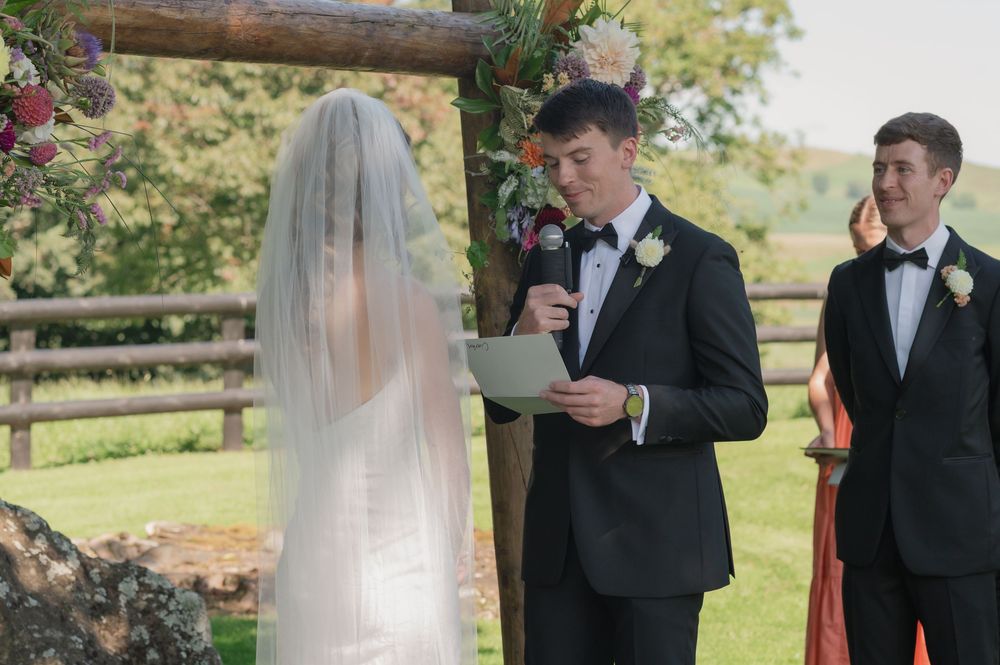 Groom reads vows to bride in white veil at outdoor wedding ceremony with wooden arch backdrop.