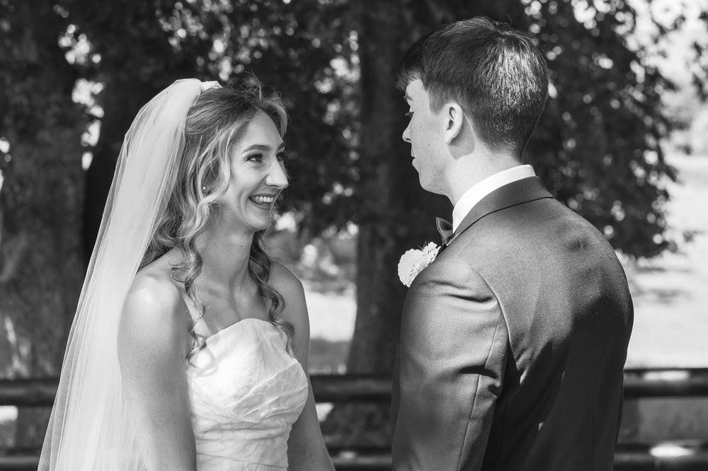 Black and white wedding photo of smiling bride in veil and groom in tuxedo sharing a romantic moment outdoors.