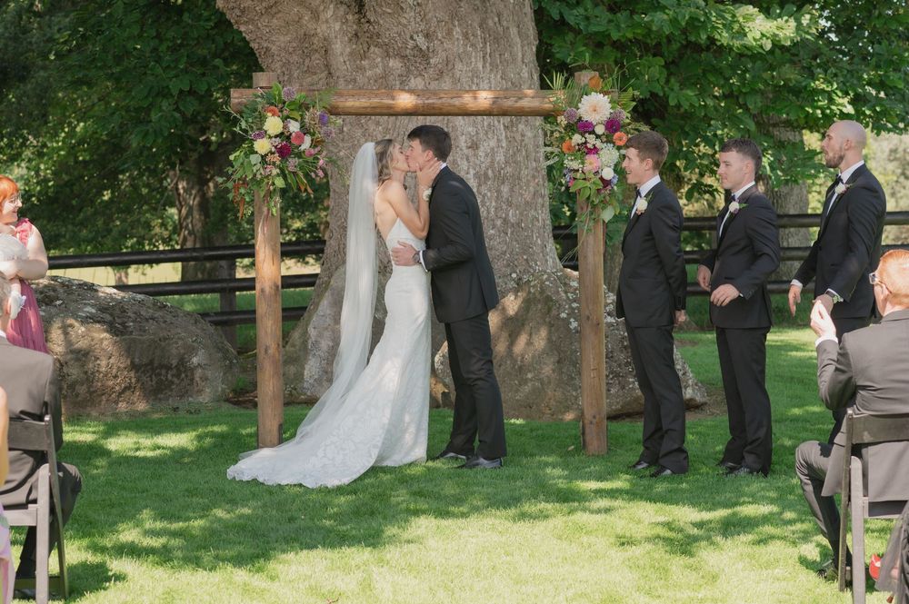 Bride and groom share first kiss under wooden arch with floral decor during outdoor wedding ceremony on green lawn.