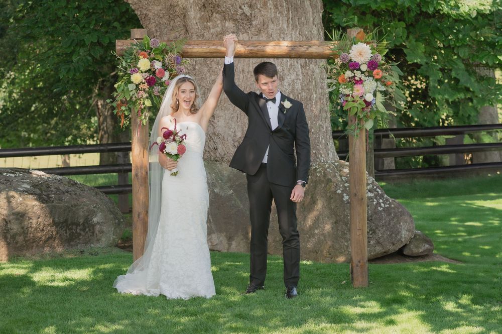 Joyful newlyweds celebrate under floral wooden arch as groom raises arm in triumph at outdoor wedding.