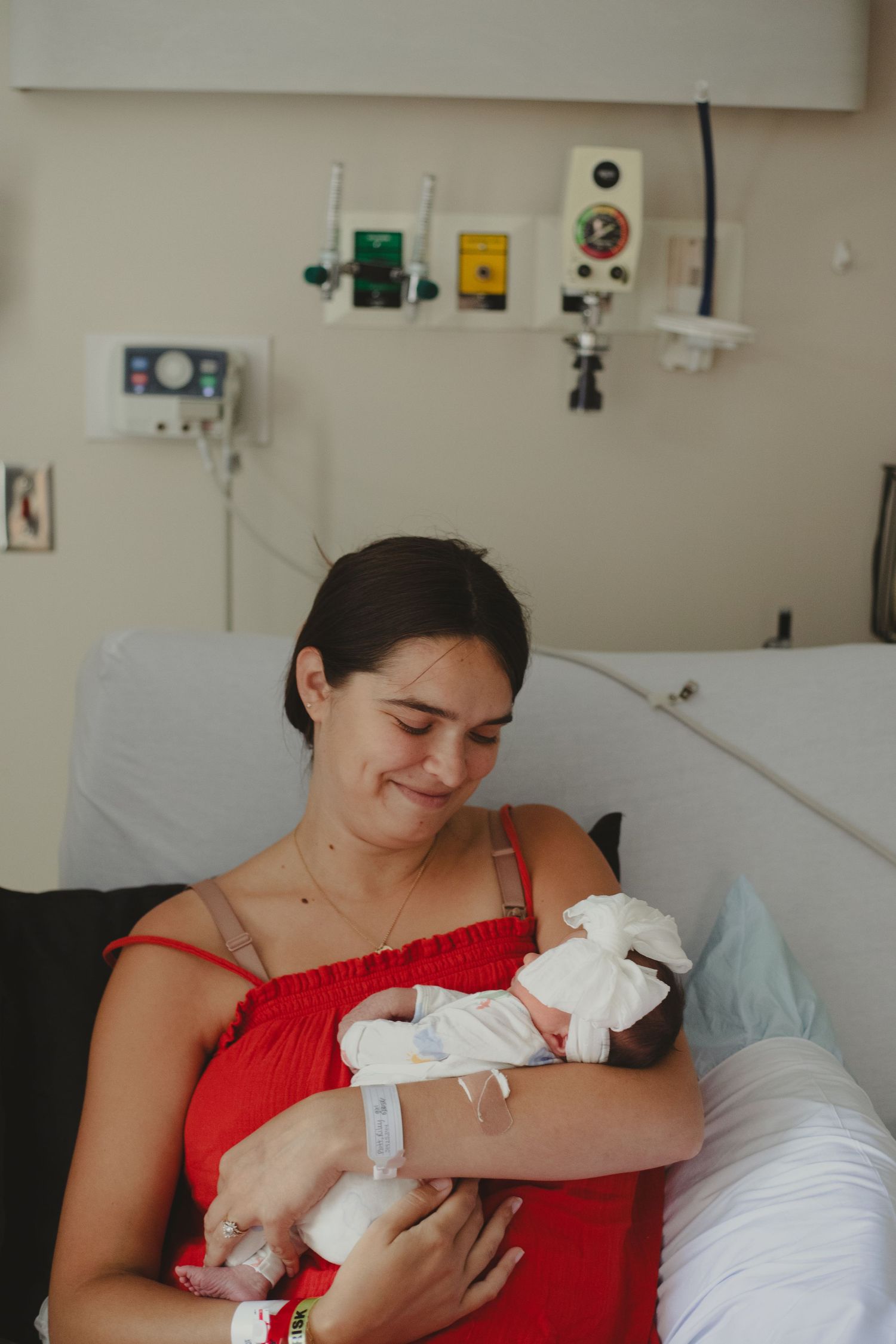 Smiling new mother in red dress cradling her newborn baby in a hospital room.